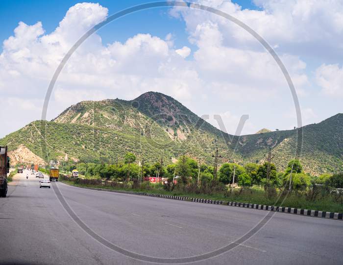 Wide Shot Of Indian Highway With Trucks, Cars, Bikes, Motorcycle And More On A Wide Asphalt Road With Green Plants Covering Hills With Clouds Of Monsoon Rains Casting Shadows Showing The Beautiful Road Routes Of Indian