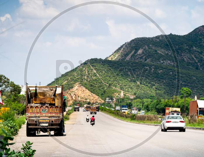 Wide Shot Of Indian Highway With Trucks Parked On Side Of Road, Cars, Bikes, Motorcycle And More On A Wide Asphalt Road With Green Plants Covering Hills With Clouds Of Monsoon Rains Casting Shadows Showing The Beautiful Road Routes Of Indian