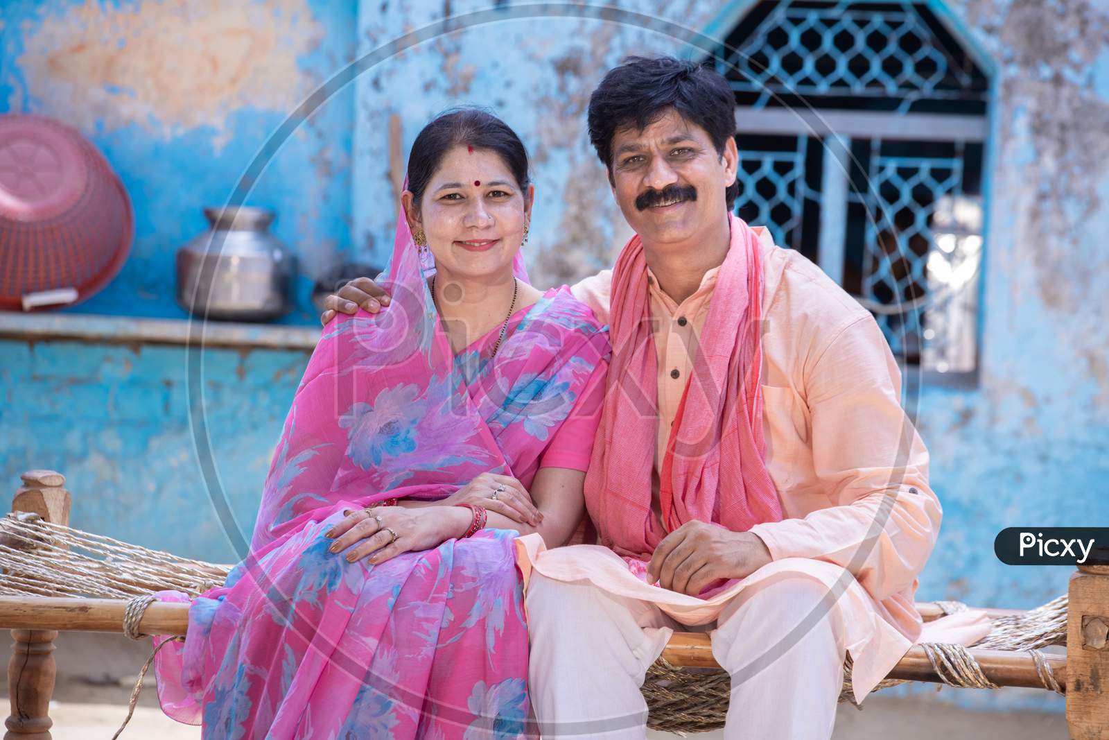 Portrait Happy Rural Indian Couple Husband Wife Wearing Traditional Outfit Sitting On Wooden Bed . Village Mustache Man And Woman Insari Smiling Looking At Camera.