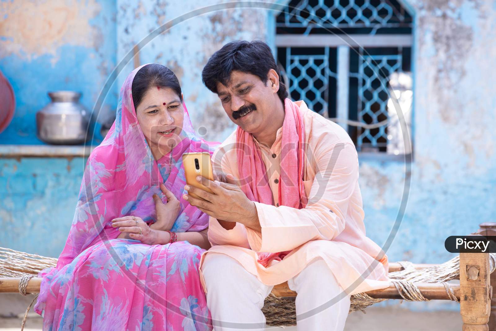 Happy Rural Indian Couple Using Smart Phone Sit On Bed. Farmer Husband Wife Watching At Mobile Screen, Man Woman Smiling.Internet Video Call,Village People Using Cellphone Technology. Digital India.