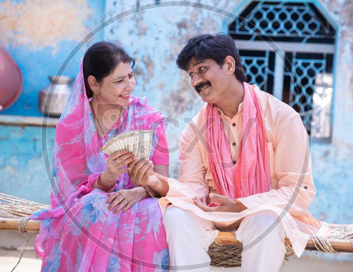 Happy Rural Woman In Sari Holding Counting Indian Rupees Note, Farmer Man Give Cash Money To Woman, Husband And Wife Sit On Bed Outside Village House. Savings And Financial Growth.