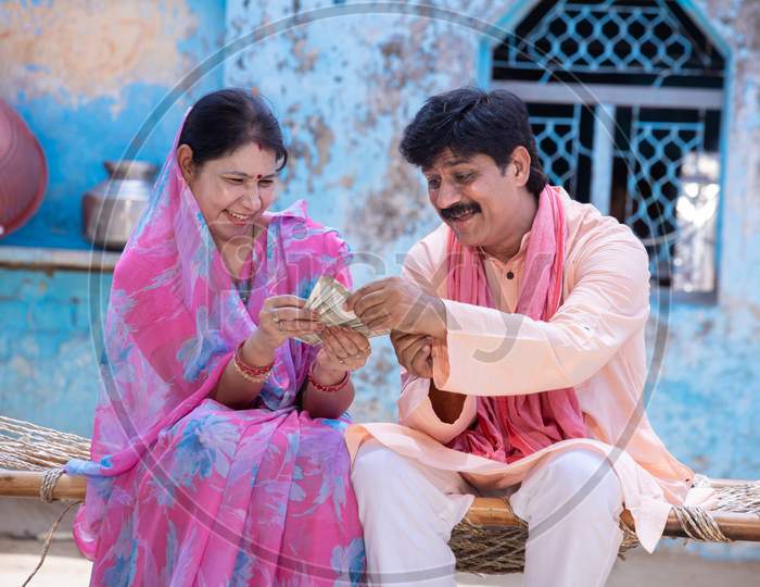 Happy Excited Rural Woman In Sari Holding Counting Indian Rupees Note, Farmer Man Give Cash Money To Woman, Surprised Husband And Wife Sit On Bed Outside Village House. Savings And Financial Growth.