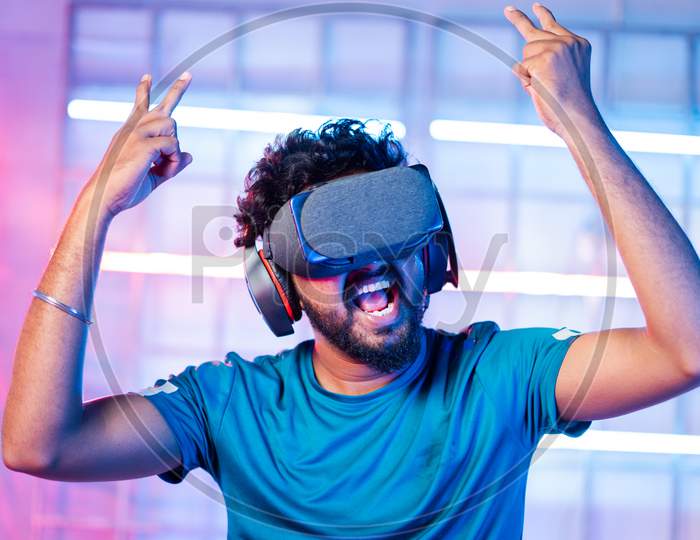 Happy Cheerful Young Man With Vr Or Virtual Reality Goggles Or Headphones Dancing On Neon Light Staged Backgound - Concept Of Modern Technological Experience.