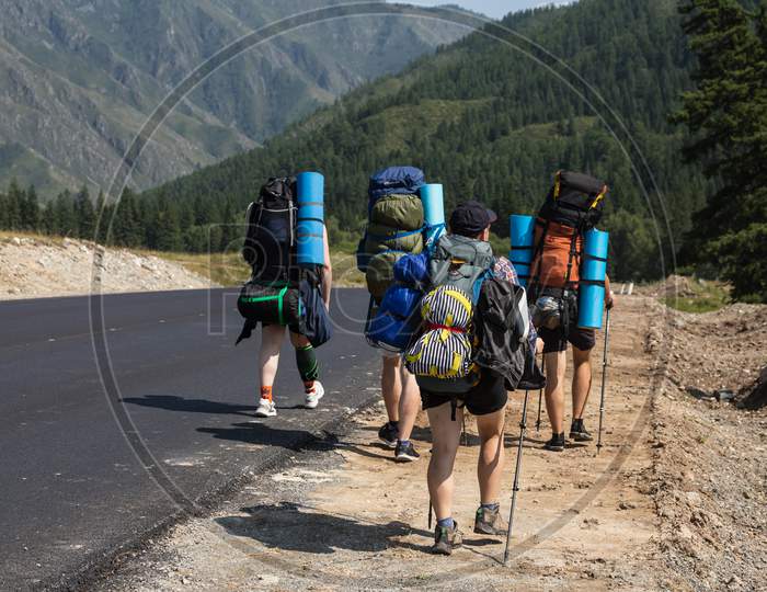 Travel Lifestyle And Survival Concept Rear View. Tourists With Backpack Go Along The Path To The Mountains In A Long Hike On A Warm Summer Day, A Back View.