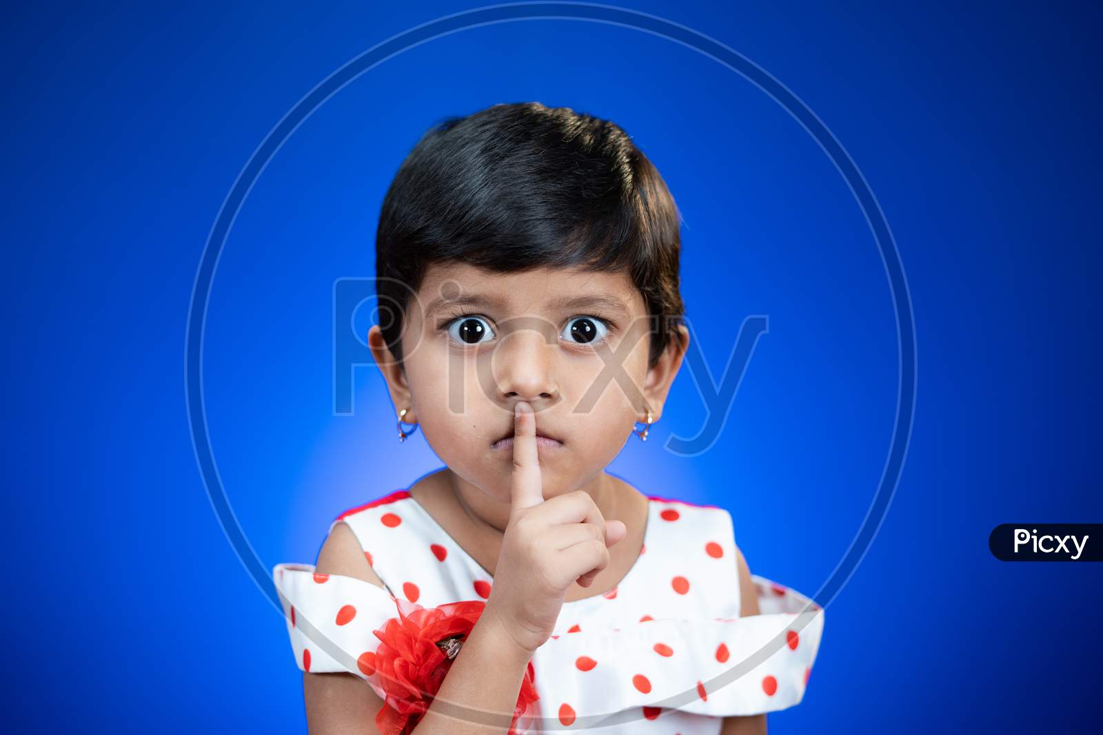 Cute Girl Kid Saying Keep Silence Quite By Placing Finger On Mouth On Blue Studio Background By Looking At Camera.