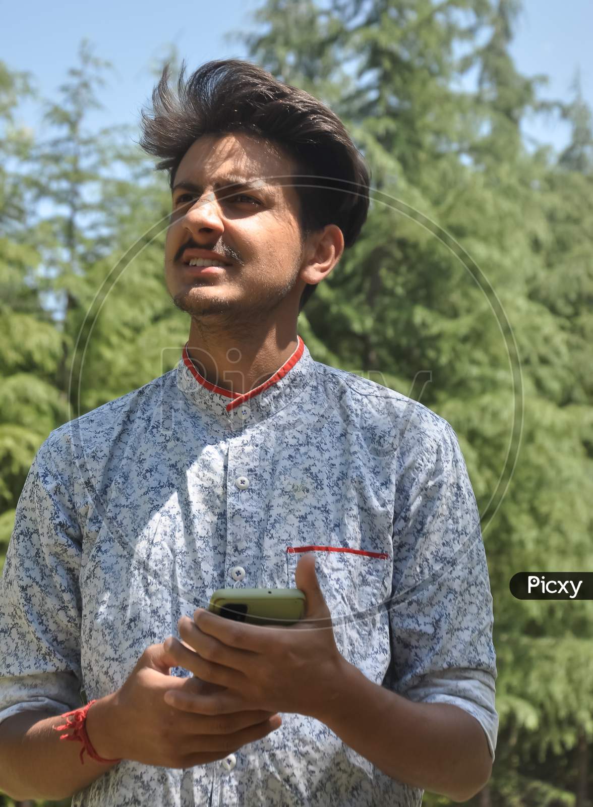 Closeup portrait of a Indian good looking young guy standing outdoor in nature and holding mobile phone in his hands with looking sideways