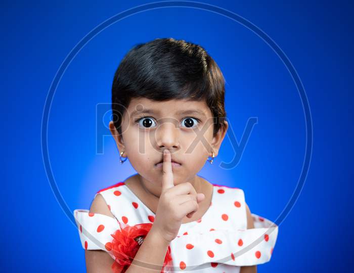 Cute Girl Kid Saying Keep Silence Quite By Placing Finger On Mouth On Blue Studio Background By Looking At Camera.
