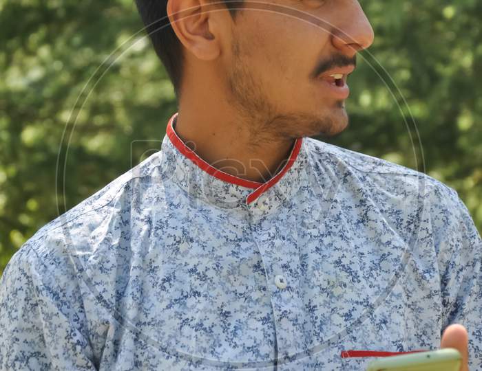 Closeup portrait of a Indian good looking young guy standing outdoor in nature and holding mobile phone in his hands with looking sideways