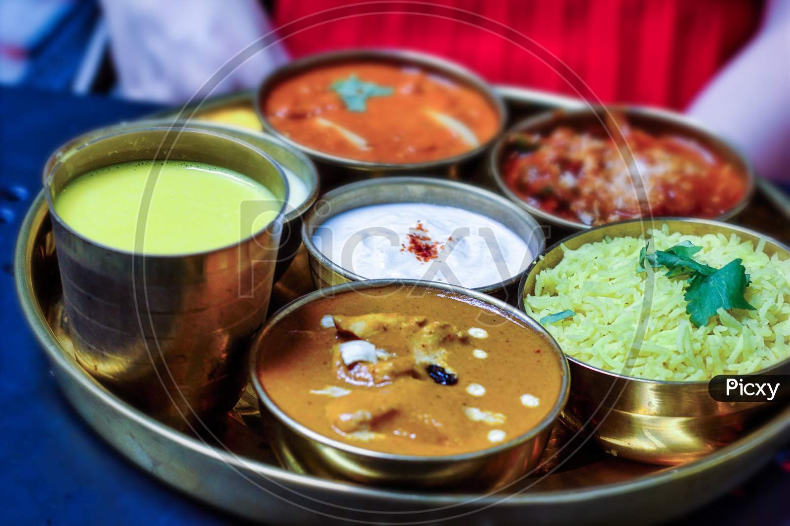 An Indian Thali Meaning Steel Plate Containing Different Food Items On A Round Platter Such As Basmati Rice, Curd Or Raita, Butter Chicken And Mango Lassi Etc I