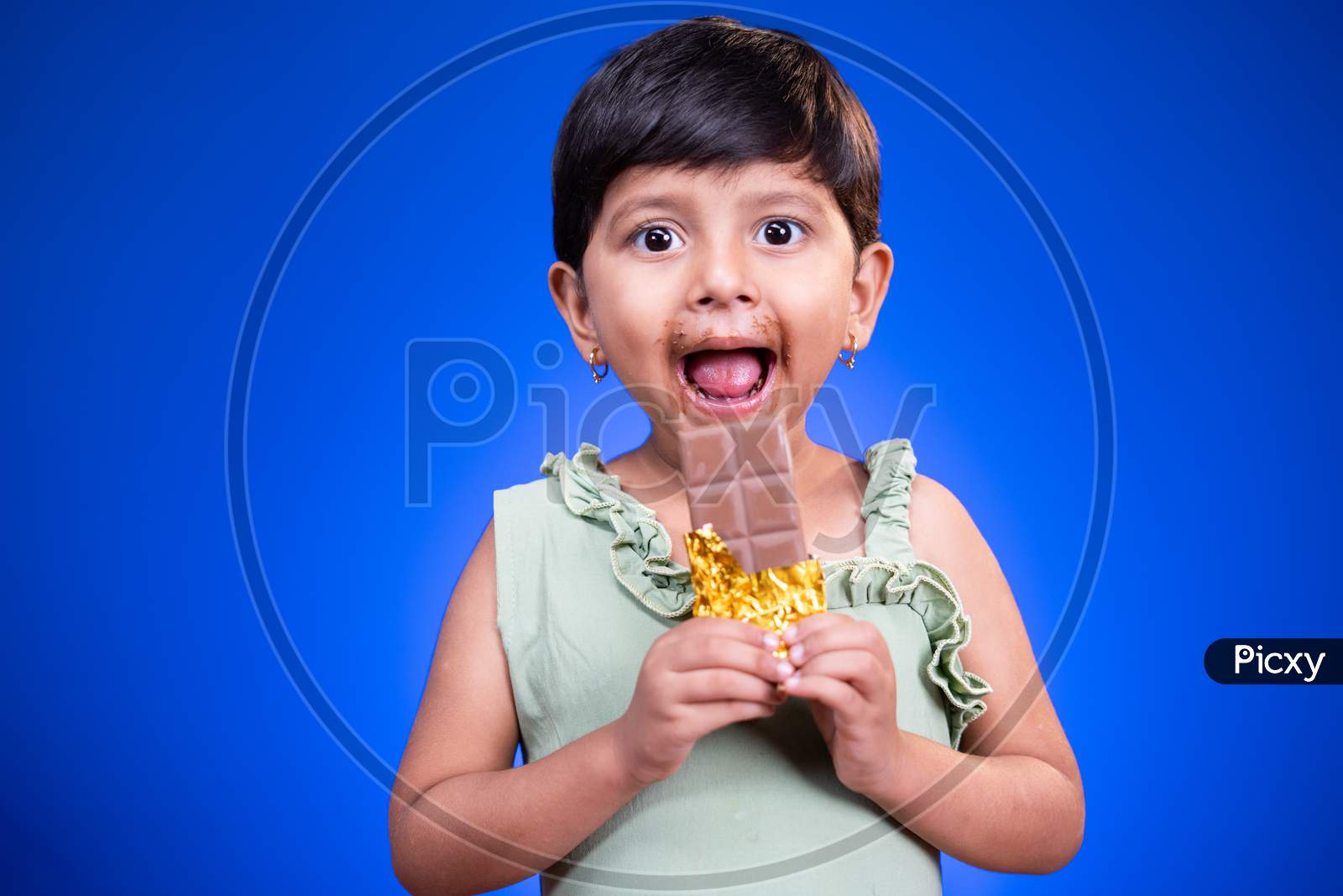 Excited Girl Kid With Large Eyes Open, Enjoying Eating Chocolate On Blue Studio Background - Concept Of Unhealthy Food Consumption And Childrens Love On Chocolate