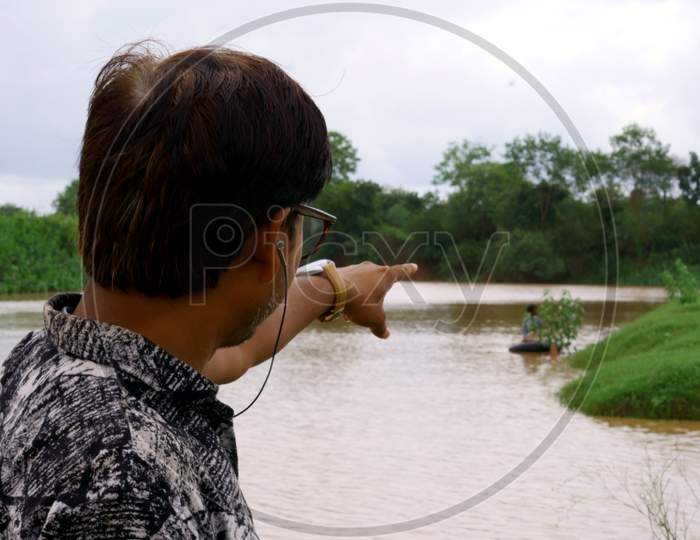 Smart Man Wearing Headphone And Specs While Pointing Finger Towards Fisherman At River Bank.