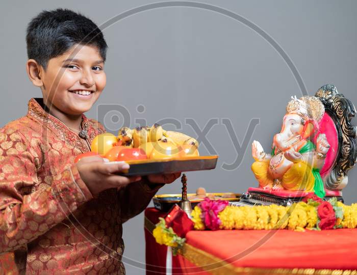 Studio Shot Of Happy Smiling Kid Offering Fruits To Lord Ganesha By Looking At Camera During Festival Celebrations.