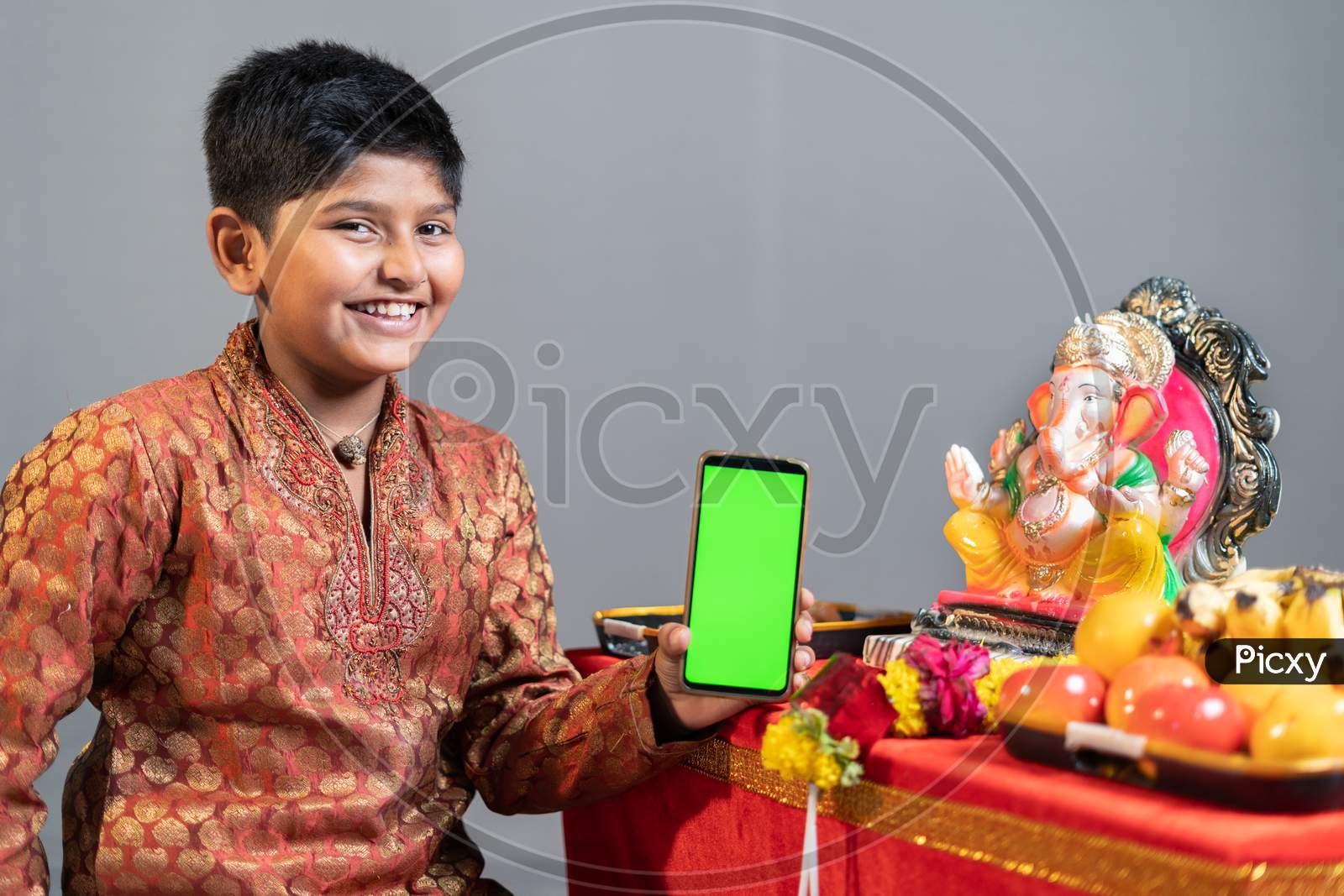 Studio Shot Of Happy Smiling Kid Holding Mobile Phone With Green Screen In Front Of Lord Ganesha Idol During Festival Celebrations.