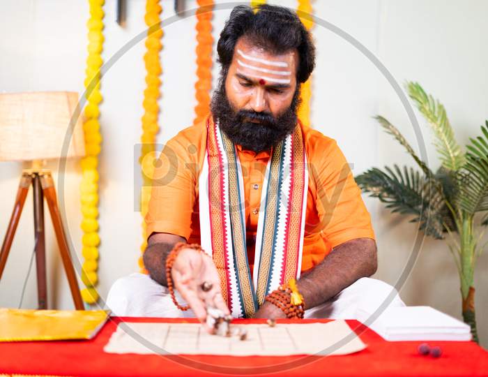 Beard Priest, Holy Astrologer Guru Or Jyotishi Placing Cowrie Shells On Chart And Counting To Predict Future By Looking Into Book - Concept Of Indian Horoscope Or Fortune Telling.