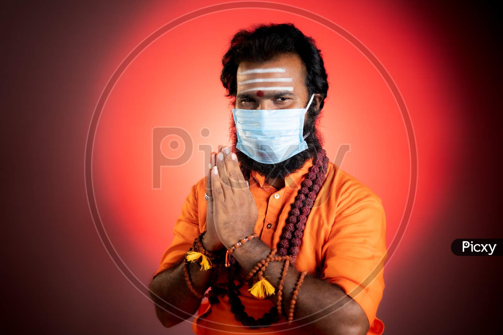 Smiling Indian God Man Or Holy Priest Doing Namaste With Medical Face Mask By Looking At Camera - Concept Showing Of Saint Or Minister Of Hindu Religion During Coronavirus Covid-19 Pandemic At Monastery.