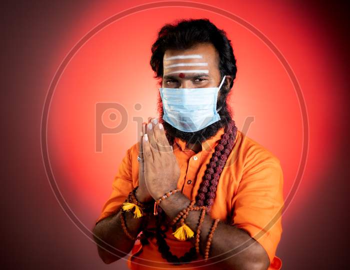 Smiling Indian God Man Or Holy Priest Doing Namaste With Medical Face Mask By Looking At Camera - Concept Showing Of Saint Or Minister Of Hindu Religion During Coronavirus Covid-19 Pandemic At Monastery.
