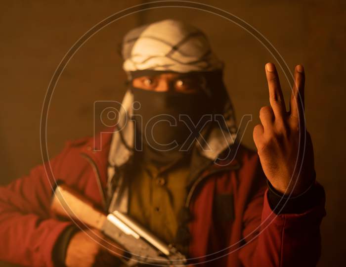 Happy Islamic Militant Or Soldier With Face Cover Showing V Sign Or Victory Gesture On Warfare With Gun In Hand