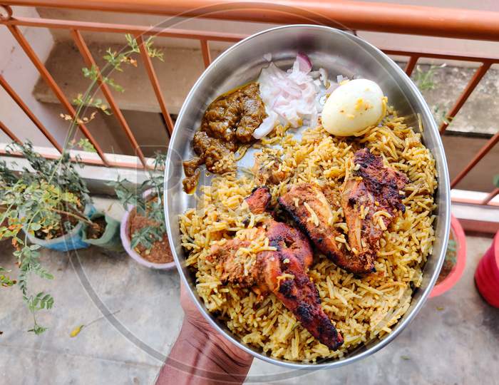 Male Hands Holding A Silver Plate Filled With Chicken Biryani, Two Tandoori Chicken, One Boiled Egg , Raitha And Brinjal Curry. A South Indian Sunday Lunch.