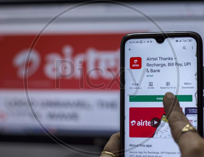 A Female Hand Holding A Android Smart Phone In Front Of A Desktop Computer And Comparing Airtel Plans.