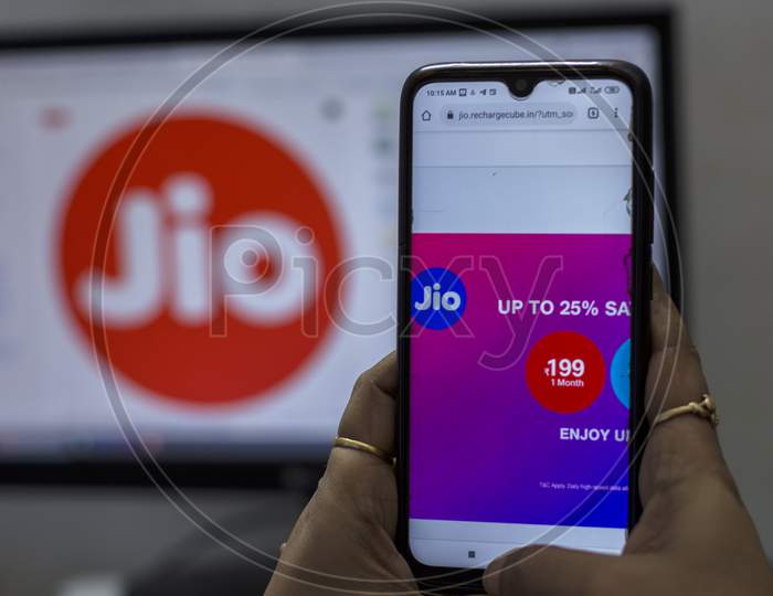 A Female Hand Holding A Android Smart Phone In Front Of A Desktop Computer And Comparing Jio Plans.