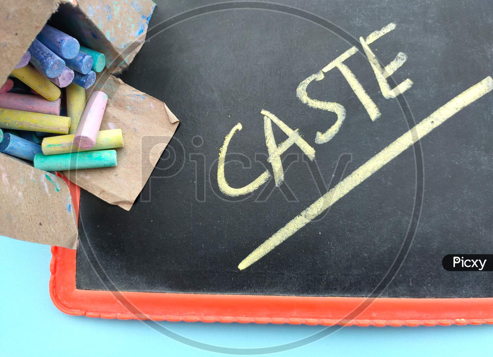 Colorful Chalk In The Box And Slate On The Table And Caste Written On It.