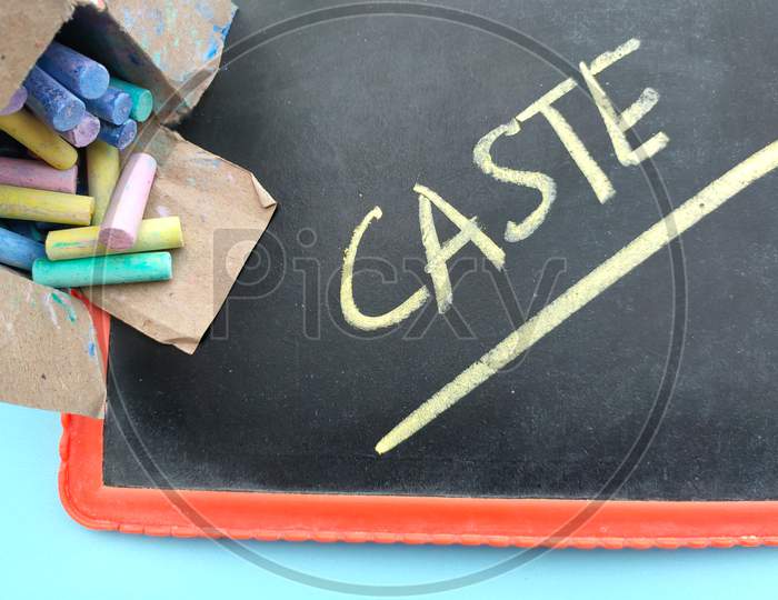 Colorful Chalk In The Box And Slate On The Table And Caste Written On It.