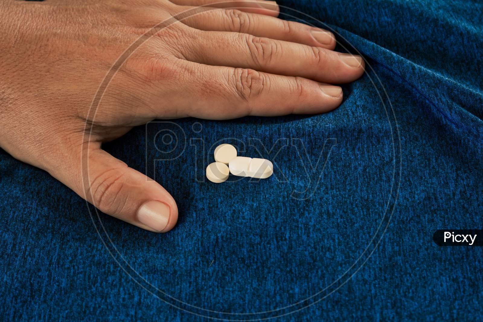 Pills On A Blue Textile Background Next To A Hand Of A Caucasian Man.