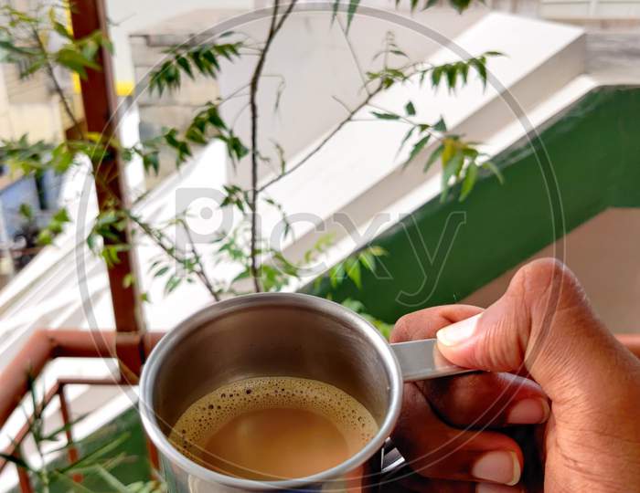 Hand Of Young Boy Holding A Silver Cup Filled With Coffee On Neem Trees Background. Evening Vibes