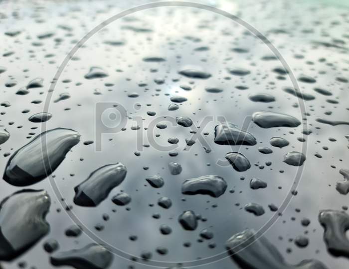 Rain Drops On A Black Metallic Car Surface In A Closeup View.
