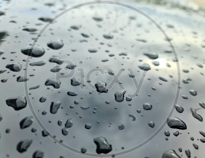 Rain Drops On A Black Metallic Car Surface In A Closeup View.