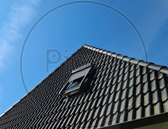 Open Roof Window In Velux Style With Black Roof Tiles And A Blue Sky Background.