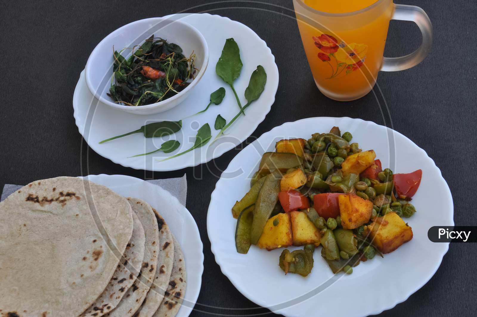 Photo of matar paneer mix veg, saag (greens) and roti (Indian bread) on white plates and juice on glass over black table