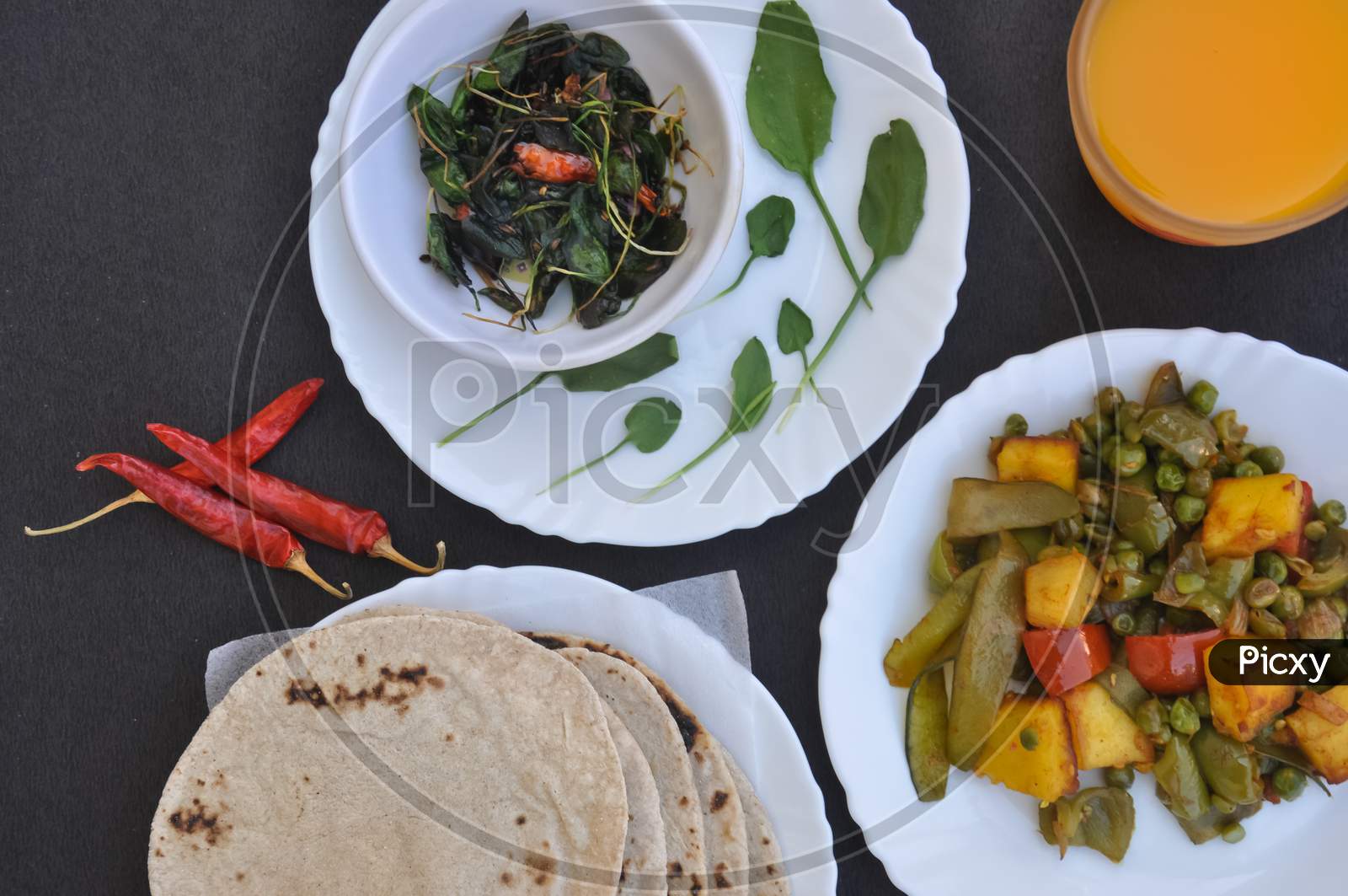 High angle of matar paneer mix veg, saag (greens) and roti (Indian bread) on white plates and juice on glass over black table