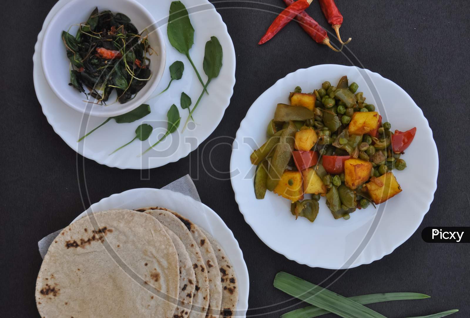 Photo of matar paneer mix veg, saag (greens) and roti (Indian bread) on white plates over black background.
