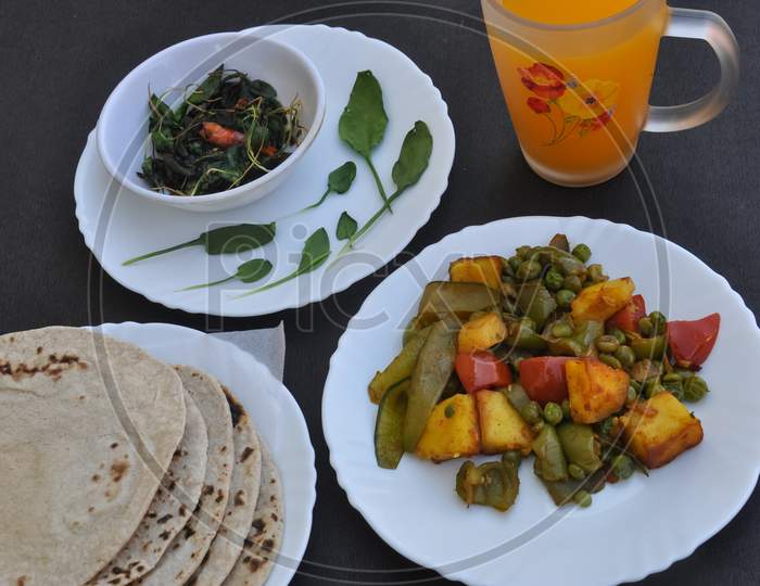 Photo of matar paneer mix veg, saag (greens) and roti (Indian bread) on white plates and juice on glass over black table