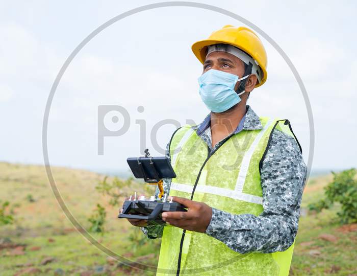 Drone Operator With Safety Helmet And Face Mask Operating Drone Using Remote Controller - Concept Of Engineer Doing Aerial Survey Using Uav During Coronavirus Covid-19 Pandemic