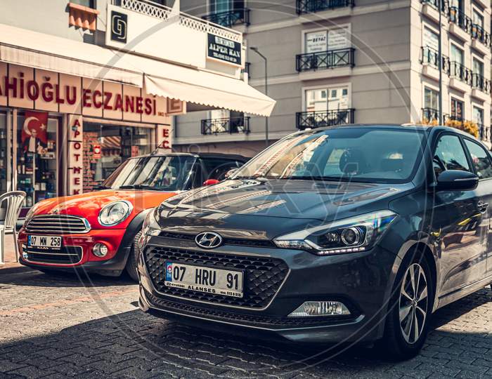 Alanya, Turkey – April 12 2021:    Gray Hyundai I20 And  Red Mini Cooper  Parked  On The Street In City Against The Backdrop Of A   Buildung,  Shops, Trees