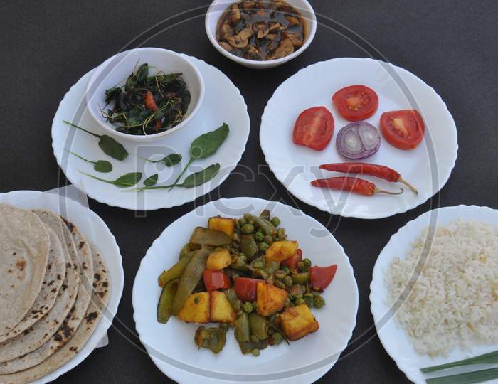 Indian Food - Mushroom soup, saag (greens), salad, chapati (Indian flat bread), matar paneer mix veg and chawal (rice) in white plates over black background