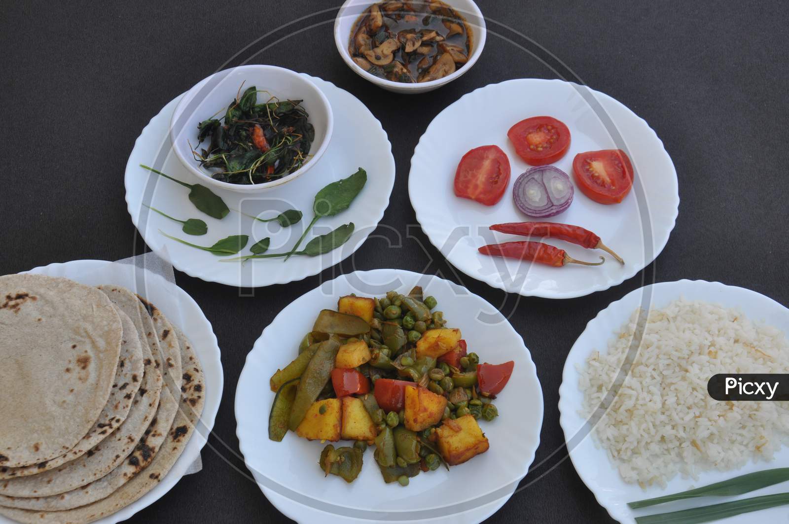 Indian Food - Mushroom soup, saag (greens), salad, chapati (Indian flat bread), matar paneer mix veg and chawal (rice) in white plates over black background