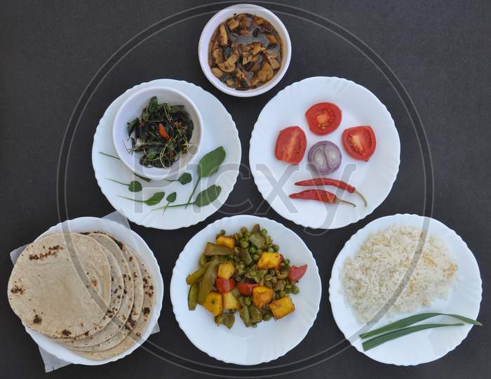 Indian Food - High angle view of mushroom soup, saag (greens), salad, roti (Indian bread), matar paneer mix veg and chawal (rice) in white plates over black background