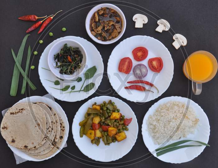 Indian Food: Overhead view of mushroom soup, saag (greens), salad, roti (Indian flat bread), matar paneer mix veg, chawal (rice) and juice over black background