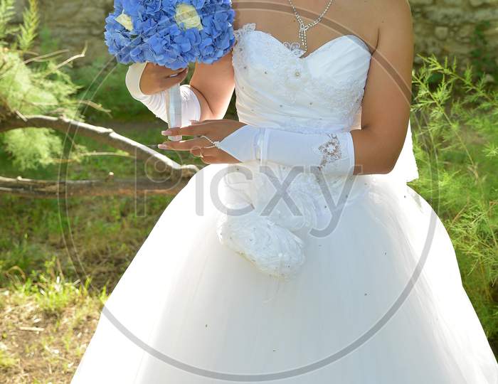 Outdoor Portrait Of Bride Dressed In White. The Bride Is Holding A Blue Flower Bouquet