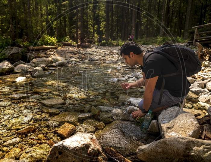 Brown Indian Arab Man With Backpack Sitting Next To Clean Water Stream Or River Drinking Water From It, Taking Break While Trekking Or Hike At Forest Creek . Adventure In Nature During Summer Day.