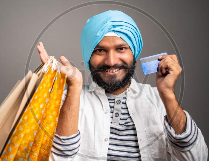 Happy Young Sikh Man With Shopping Bag And Credit Card In Hand Looking Into Camera On Studio Background - Concpet Of Online E-Commerce Festival Shopping.