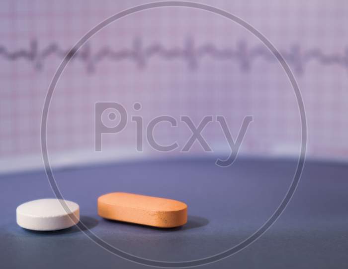 Two Pills On A Neutral Blue Background. An Ekg Trace Is Visible In The Unfocused Background.