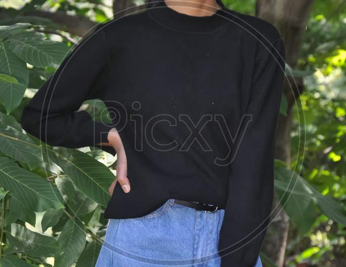 Portrait of a charming young girl standing outside and wearing black sweatshirt and light blue jean and posing with her hand on hips and looking at camera
