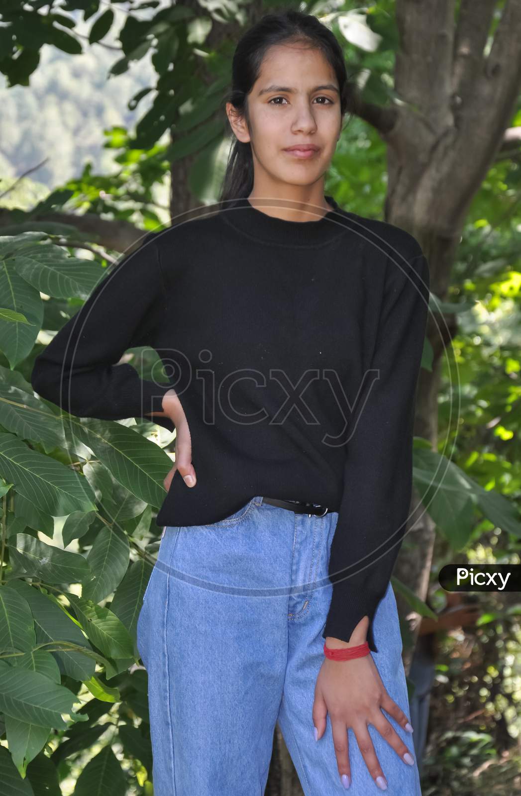Portrait of a charming young girl standing outside and wearing black sweatshirt and light blue jean and posing with her hand on hips and looking at camera