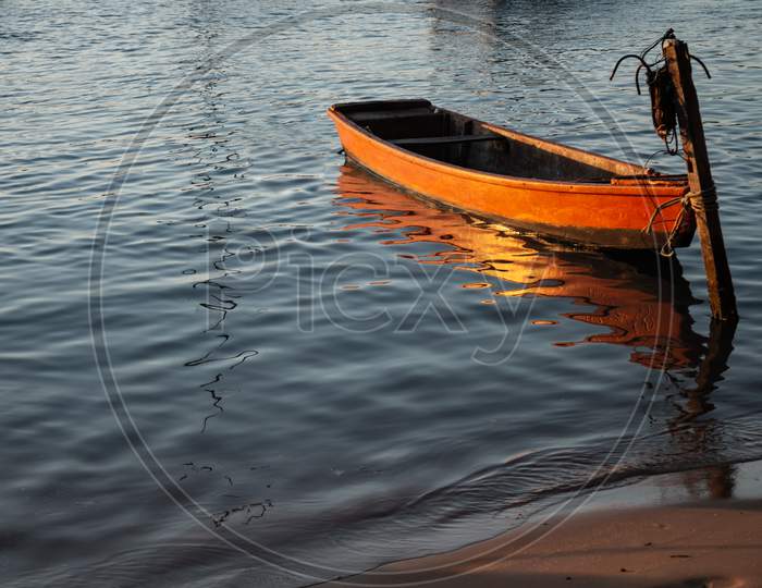 Small Orange Wooden Boat Moored To A Pole At The Edge Of The Lake During Sunset. Small Boat For Fishermen.