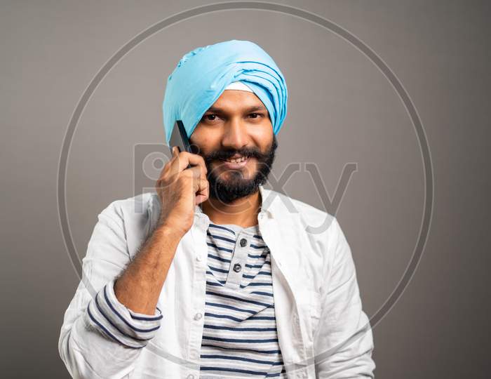 Happy Smiling Indian Sikh Man Busy Talking On Mobile Phone On Studio Background By Looking Camera - Concept Of Communication, Network And Positive Emotions