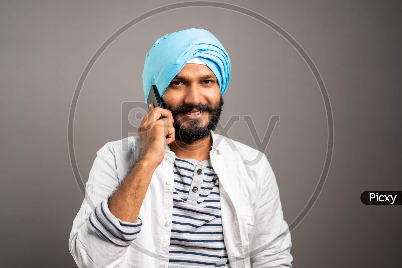 Happy Smiling Indian Sikh Man Busy Talking On Mobile Phone On Studio Background By Looking Camera - Concept Of Communication, Network And Positive Emotions